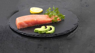 Avokado dilimli somon filetosu siyah taş tahtada limon ve otlar.