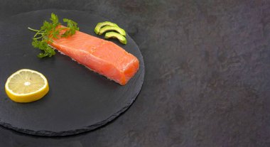 Avokado dilimli somon filetosu siyah taş tahtada limon ve otlar.