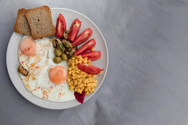 Kızarmış yumurta, mısır domatesli zeytin ve beyaz tabakta servis edilen ekmek.