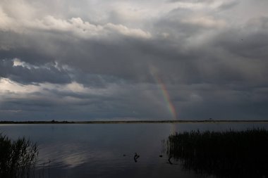 Dramatik fırtına bulutlarıyla sakin göl manzarası ve su üzerinde ince gökkuşağı yansıması