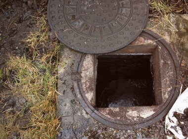 Islak zeminde kısmen kaymış metal kaplı, çimenler ve çamurla çevrili, açık hava ortamında karanlık yeraltı atık su kuyusunu gösteren kanalizasyon bacası.