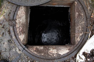 Islak zeminde kısmen kaymış metal kaplı, çimenler ve çamurla çevrili, açık hava ortamında karanlık yeraltı atık su kuyusunu gösteren kanalizasyon bacası.