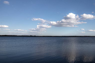 Durgun su yüzeyi mavi gökyüzünün altında beyaz bulutlar ve uzak ufuklarla