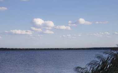 Mavi gökyüzünün altında, beyaz bulutlar ve uzak orman ufkuyla sakin bir göl.