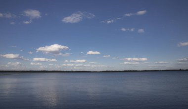Durgun su yüzeyi mavi gökyüzünün altında beyaz bulutlar ve uzak ufuklarla