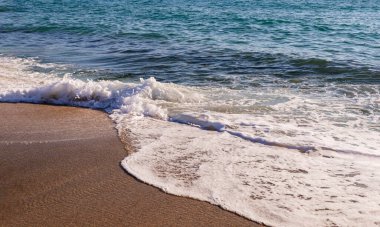 Deniz dalgaları kumlu sahile vuruyor. Su yüzeyinde parlak güneş ışığı yansımaları var.