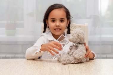 a dark-skinned girl sits at a table in a white shirt with pigtails and gives a toy bear water to drink