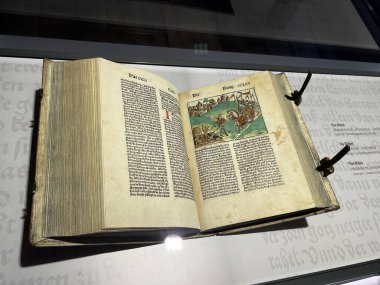 Açık bir erken basılmış Alman İncili, siyah metin ve sağ sayfada bir savaşın ya da olayın ahşap kesim illüstrasyonunu gösteriyor, vitrin vitrininde metal kıskaçlarla korunuyor..