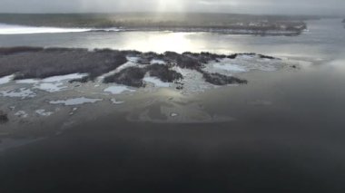 Kamera kışın başında bir kuadrokopterle nehri yakalar. Nehirde biraz buz göründü bile. Nehrin ortasında bir ada. Güneş, suyun yüzeyine yansıyor. Nehrin kıyısında bir orman var.