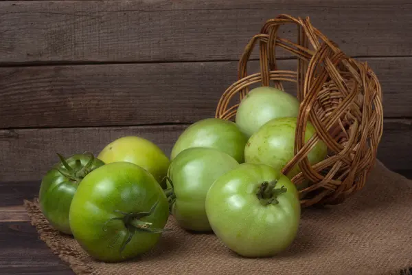 Unreife grüne Tomaten im Weidenkorb auf hölzernem Hintergrund und Sacktuch — Stockfoto