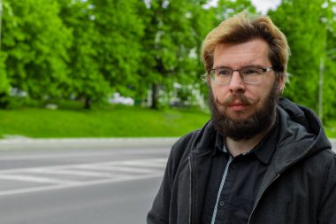 Bıyıklı, beyaz yüzlü, orta yaşlı insan portresi olan sakallı bir adam. Sokakta yeşil ağaçlı, bahar manzaralı.