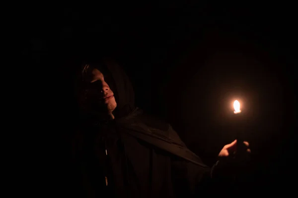 mysterious man in robe with hood and candle in hand ready for scary story telling before night, soft focus photography on Halloween thematic with black background space for copy or text