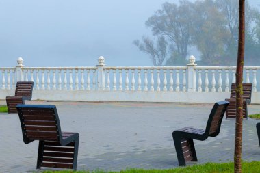 Puslu, sıcacık bir sahil kenarı, klasik mimari fayans mermer çit, sabah sisli arka plan ve burada olmayan insanlar için post modernizm tarzı sandalyeler.