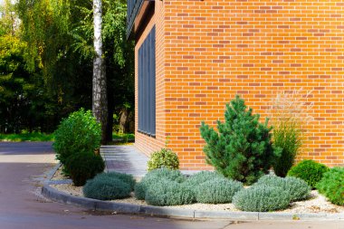 Çağdaş bir bina cephesinde turuncu tuğla ve koyu renkli pencereler, iyi korunmuş bir bahçe ortamında çeşitli süs bitkileri ve ağaçlarla tamamlanmış.