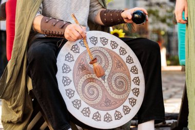 Ortaçağ tarzı giyinmiş bir kişi elinde siyah bir kristal top tutarken büyük ve süslü bir davul çalar. Bu da ruhani veya törensel bir bağlam olduğunu gösterir.