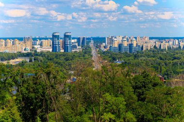 Panoramik bir manzarada yoğun bir şehir manzarası, bol yeşil yapraklarla çevrili ve ufka doğru uzanan yoğun bir otoyol, parçalı bulutlu bir gökyüzü altında sergilenmektedir.