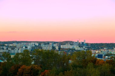 Akşam gökyüzünde pembe ve turuncu renklerin yumuşak bir gradyanı altında genişleyen kentsel manzara, manzarayı çevreleyen yoğun yeşilliklerle gözler önüne seriliyor.
