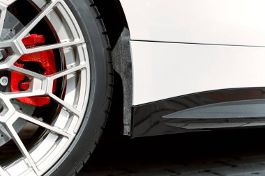 Beyaz lüks bir araçta gümüş alaşımlı bir tekerlek, canlı kırmızı fren kalibreli ve aerodinamik bir yan eteğin karmaşık tasarımını gösteren detaylı bir manzara.
