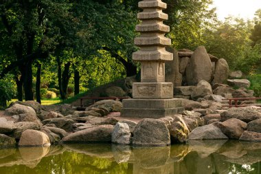 Yıpranmış taş bir pagoda, sakin bir göletin yanındaki kayalık bir arazide duruyor. Etrafı canlı ağaçlar ve yapraklarla çevrili. Huzurlu bir atmosfer yaratıyor.