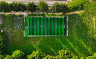 Yukarıdan çekilen insansız hava aracı, yeşil çizgili desenli, yemyeşil bir futbol sahası yakalıyor. Etrafı çim ve ağaçlarla çevrili, futbol oynayan küçük bir grup insan ve spor yapan bir grup insan görülüyor.
