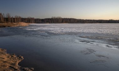 Baharın erken saatlerinde gün batımında gölün kumlu kıyıları. Ufukta ağaçlar ve sıcak alacakaranlık gökyüzü olan buzlu su yüzeyi..