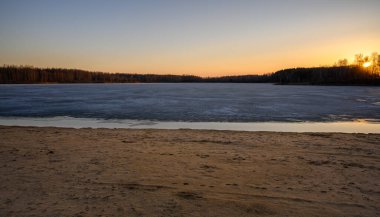 Baharın başlarında bir gölün kıyısında renkli bir günbatımı. Güneş arka plandaki ağaçların arkasında saklanıyor. Yüzeyin yarısı buzla kaplı..