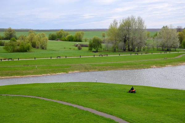 Yeşil kıyılarında bir nehir ve çim biçme makinesi olan güzel bir park. İlkbahar manzarası bulutlu ve mavi gökyüzü kırsalda eğlence ve dinlenme için..