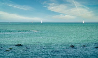 Mavi bulutlu gökyüzüne ve öndeki dalgalı su taşlarına karşı denizde yelken açan yatlar. Yaz tatili etkinliği, eğlence ve turizm konsepti..