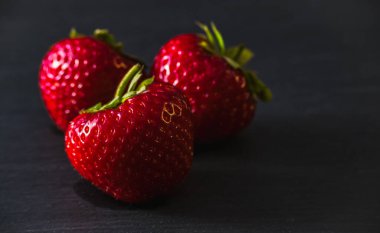 Taze olgun çilekler ya da Fragaria Ananassa koyu bir arka planda. Yeşil saplı üç kırmızı dut ve yumuşak gölgeli yapraklar. Diyet, doğal ürünler, hasat, beslenme, kapalı alan sanat dekorasyonları.