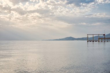 Scenic seascape view with a pier at a side at sunset in springtime. Wavy surface, overcast sky with sun rays passing through the clouds. Travel, serenity, rest, recreation, scenic destinations.