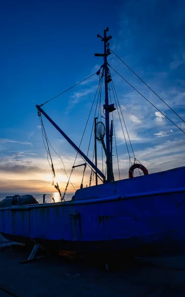 Gün batımında deniz kıyısında paslı mavi bir balıkçı teknesi. Sarı güneş ışınları su yüzeyine yansıyor. Balıkçılık, ulaşım, seyahat ve deniz manzarası kavramı.