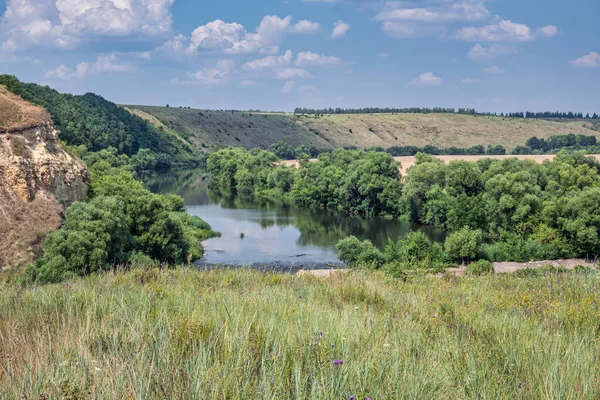 A turn of the river with high steep stone banks, covered with lush green trees and bushes. Countryside landscape on a summertime day.