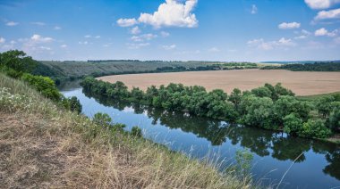 Yüksek sarp kıyıları olan, yemyeşil ağaçlar ve çalılarla kaplanmış bir nehir kıvrımı. Yansımalı pürüzsüz su yüzeyi. Bir yaz gününde kırsal alan.