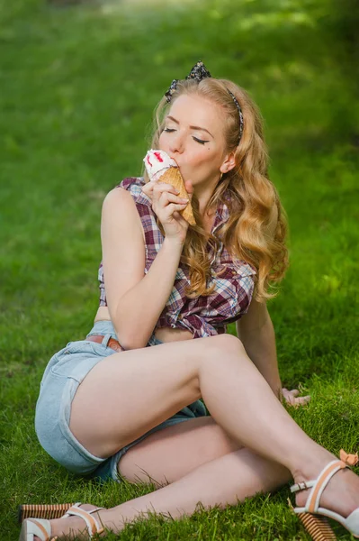 Sexy chica rubia en pantalones cortos lamiendo helado sentado en la hierba al aire libre — Foto de Stock