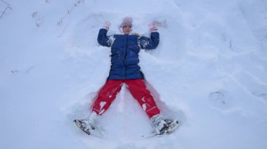 Çocuk karda yatarken bir kar meleği yapıyor ve gülümsüyor. Yavaş çekim.