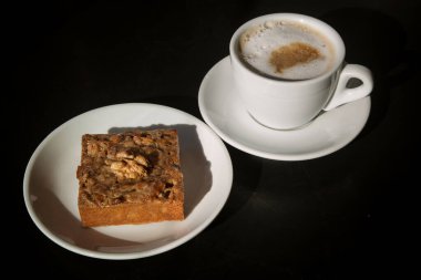 kapuçino bardağının yanındaki beyaz tabakta cevizli kek, güneşli bir fotoğraf.