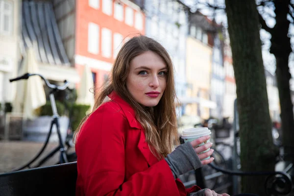 Güzel genç Avrupalı bir kız kışın bir bankta oturarak bir fincan kahve tutar. Hava soğuk..