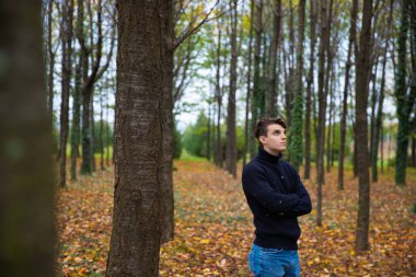 A young man with folded arms looks at the sky and stands in an autumn forest,the leaves are yellow and have fallen from the trees,he wears a wool sweater.Pensive and thoughtful male.Loneliness concept