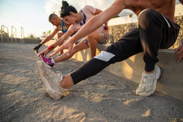 Üç kişiden oluşan bir grup, iki erkek ve bir kadın, biraz esneme egzersizi yapıyorlar, oturuyorlar ve elleriyle ayaklarına dokunuyorlar, açık havada spor salonunda ısınma egzersizi yapıyorlar.