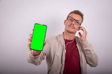 Genç çekici adam kulaklıkla müzik dinliyor, akıllı telefon ekranını gösteriyor, logosunu takmak için yeşil ekran, stüdyo fotoğrafı..