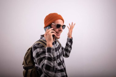 Şık bir adam telefon eder, ekose gömlek, güneş gözlüğü ve şapka takar, sırt çantalı bir hipster, stüdyoda beyaz arka planda çekilmiş bir fotoğraf.