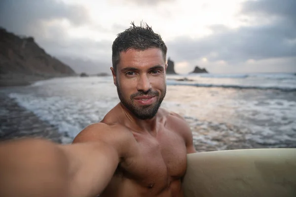 Surfer takes a selfie at the beach during sunset, he holds his surfboard and is smiling. Young attractive man at the beach - joyful person practicing extreme sport concept