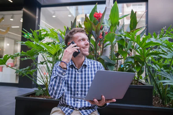 Millennial businessman talks on the phone while holding up his laptop. Smiling successful person at work - elegant caucasian man during a phone call