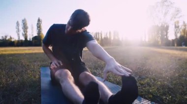 Parkta uzanan, kolları açık ayak parmaklarına uzanan genç bir adam.