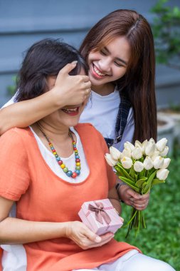 Gülümseyen annelerin gözlerini kapayan ve elinde çiçeklerle gezen kız mutlu ebeveyn hediye kutusuna sürpriz yapıyor. Anne doğum günü konsepti.