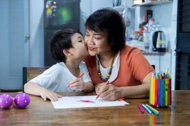 Torun evde resim çizerken büyükanneyi öpüyor. Torunu olan büyükannesi evde çalışıyor.