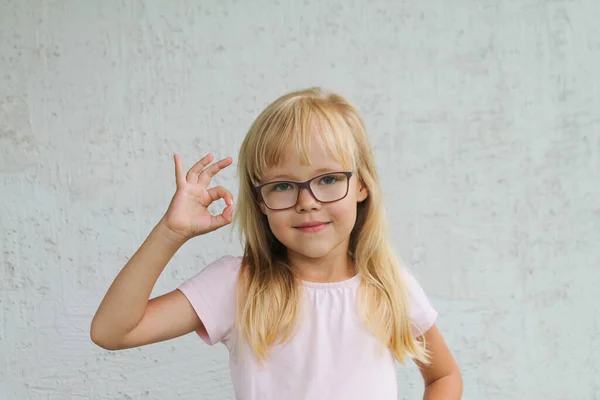 Une petite blonde avec des lunettes fait un geste OK avec un t-shirt à la menthe sur un fond gris — Photo