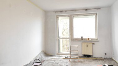 Tadilat sırasında yeni boyanmış beyaz duvarlar, merdiven, plastik zemin koruması ve pencereli boş bir oda. Ev geliştirme, apartman yenileme ve fotokopi alanı ile iç yenileme kavramı.