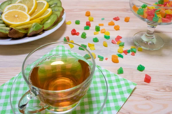 Bir bardak çay, çok renkli meyveler, beyaz ve yeşil peçeteli ahşap arka planda limon ve kivi dilimleri.