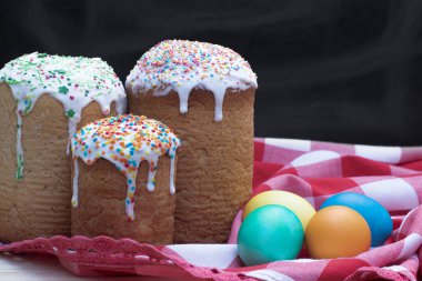 Üç fırınlanmış Paskalya pastası üzerine şeker serpiştirilmiş, dört renkli tavuk yumurtası kırmızı ve beyaz bir bez ve siyah bir arka plan, yakın plan.